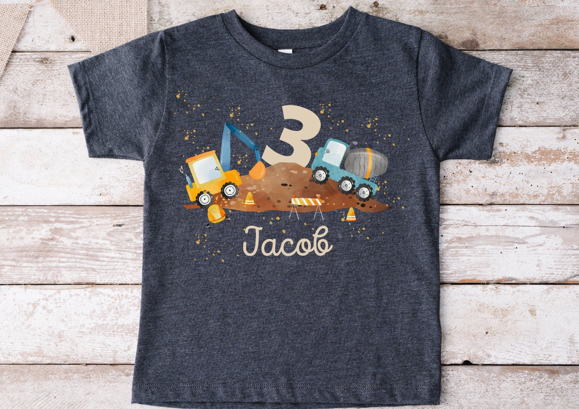 Dunkelgraues Kinder-T-Shirt mit Baustellen-Bügelbild, personalisiert mit Name Wunschname und Zahl Wunschzahl, illustriert mit Bagger, Betonmischer und Baustellen-Zubehör.